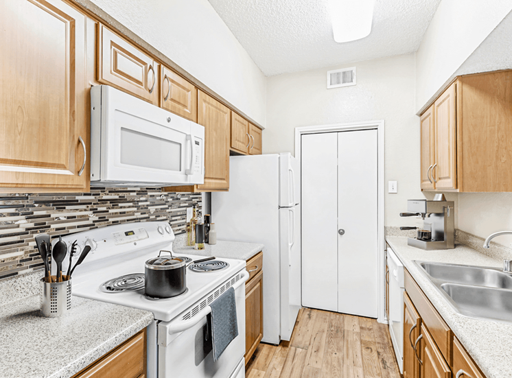 Virtually staged kitchen with white appliances, light wood cabinetry, wood style flooring, granite style countertops, double basin sink, coffee maker, kitchen utensils and pot on stove
